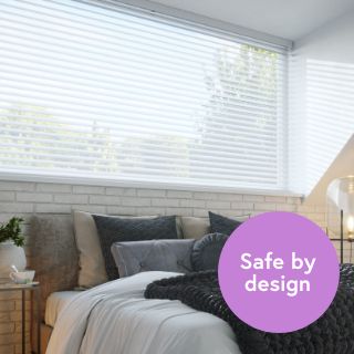 An image of a bedroom with a large window blind the bed. There are white wooden blinds installed.